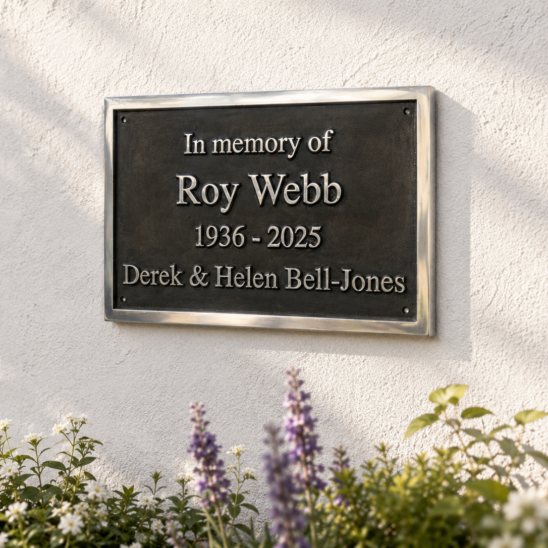 A rectangular black memorial plaque with silver lettering mounted on a white stucco wall, surrounded by greenery and flowers. The plaque reads: 'In memory of Roy Webb 1936 - 2025 Derek & Helen Bell-Jones'.