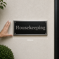 A rectangular black house number plaque with silver lettering reading 'Housekeeping' is mounted on a beige stucco wall next to a black door. A hand points to the plaque, and a dimension label '350mm' appears below it.