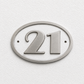 A silver oval house number plaque displaying the number 21, mounted on a textured white wall.