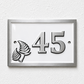 A rectangular house number plaque with a silver frame and white face, featuring the number 45 in raised silver lettering and a silver fern motif on the left, mounted on a textured white wall.