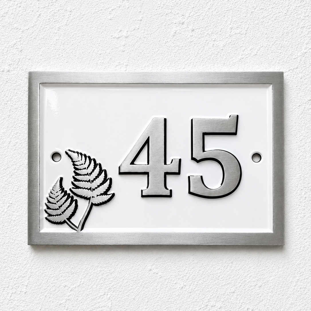 A rectangular house number plaque with a silver frame and white face, featuring the number 45 in raised silver lettering and a silver fern motif on the left, mounted on a textured white wall.