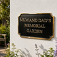 A black rectangular memorial garden plaque with gold lettering and a gold border, mounted on a white stucco wall in a garden setting.