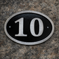 Cast aluminium oval hotel room number sign with raised numbers and black background mounted on wall
