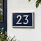 A modern house number plaque reading '23' in raised silver numerals on a dark textured blue background, mounted on a white stucco exterior wall with green foliage visible.