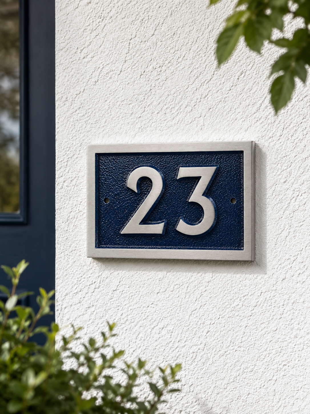A modern house number plaque reading '23' in raised silver numerals on a dark textured blue background, mounted on a white stucco exterior wall with green foliage visible.