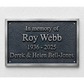 A rectangular memorial plaque mounted on a textured white wall. The plaque has a dark textured center with raised silver lettering that reads 'In memory of Roy Webb 1936 - 2025 Derek & Helen Bell-Jones' and is framed by a brushed silver border.