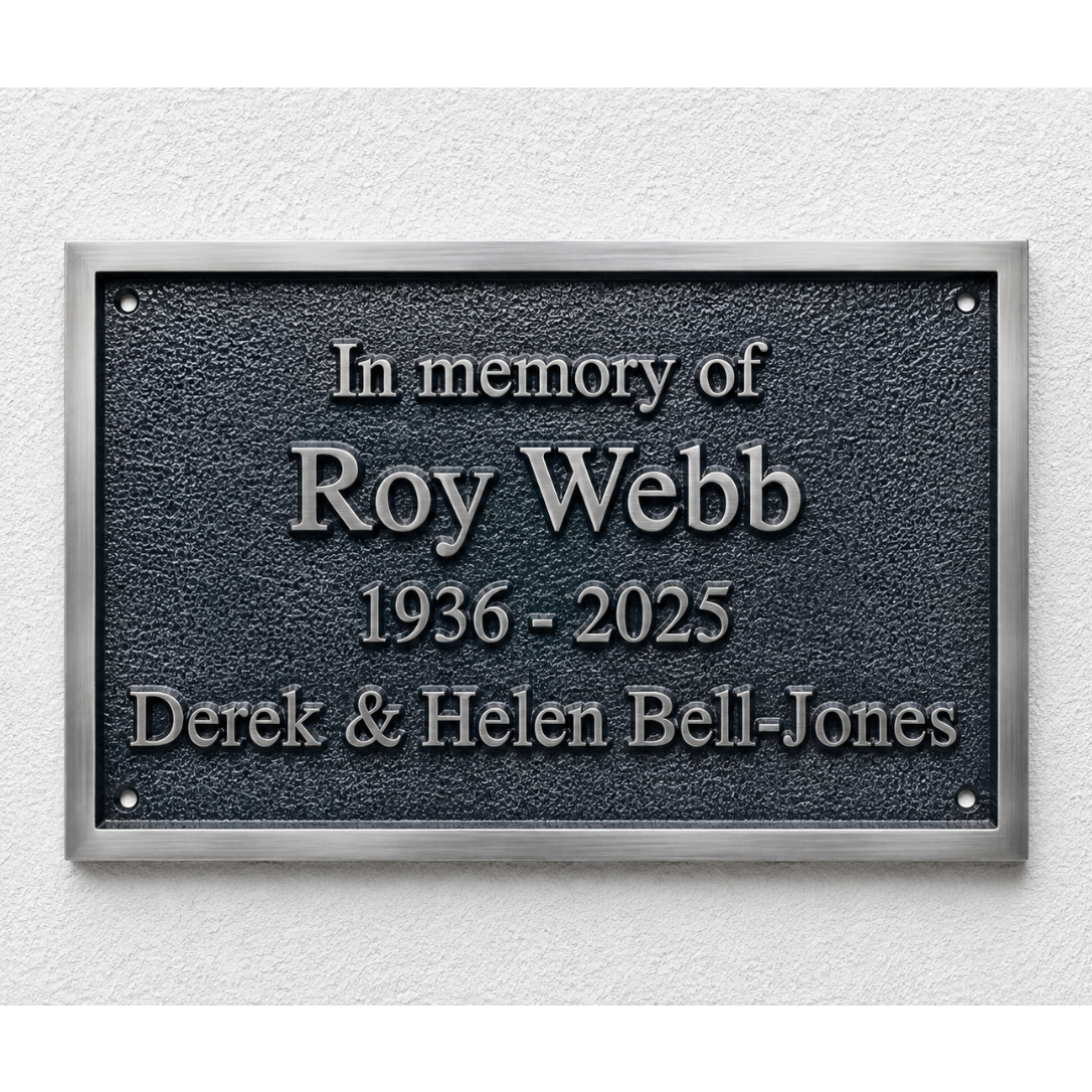 A rectangular memorial plaque mounted on a textured white wall. The plaque has a dark textured center with raised silver lettering that reads 'In memory of Roy Webb 1936 - 2025 Derek & Helen Bell-Jones' and is framed by a brushed silver border.
