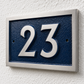 A modern house number plaque featuring the numbers '23' in raised silver lettering on a dark textured blue background, mounted on a white textured stucco wall.