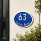 An oval blue and white house number sign reading '63 GRIFFIN ROAD' mounted on a textured white stucco wall, with green foliage partially framing the scene.
