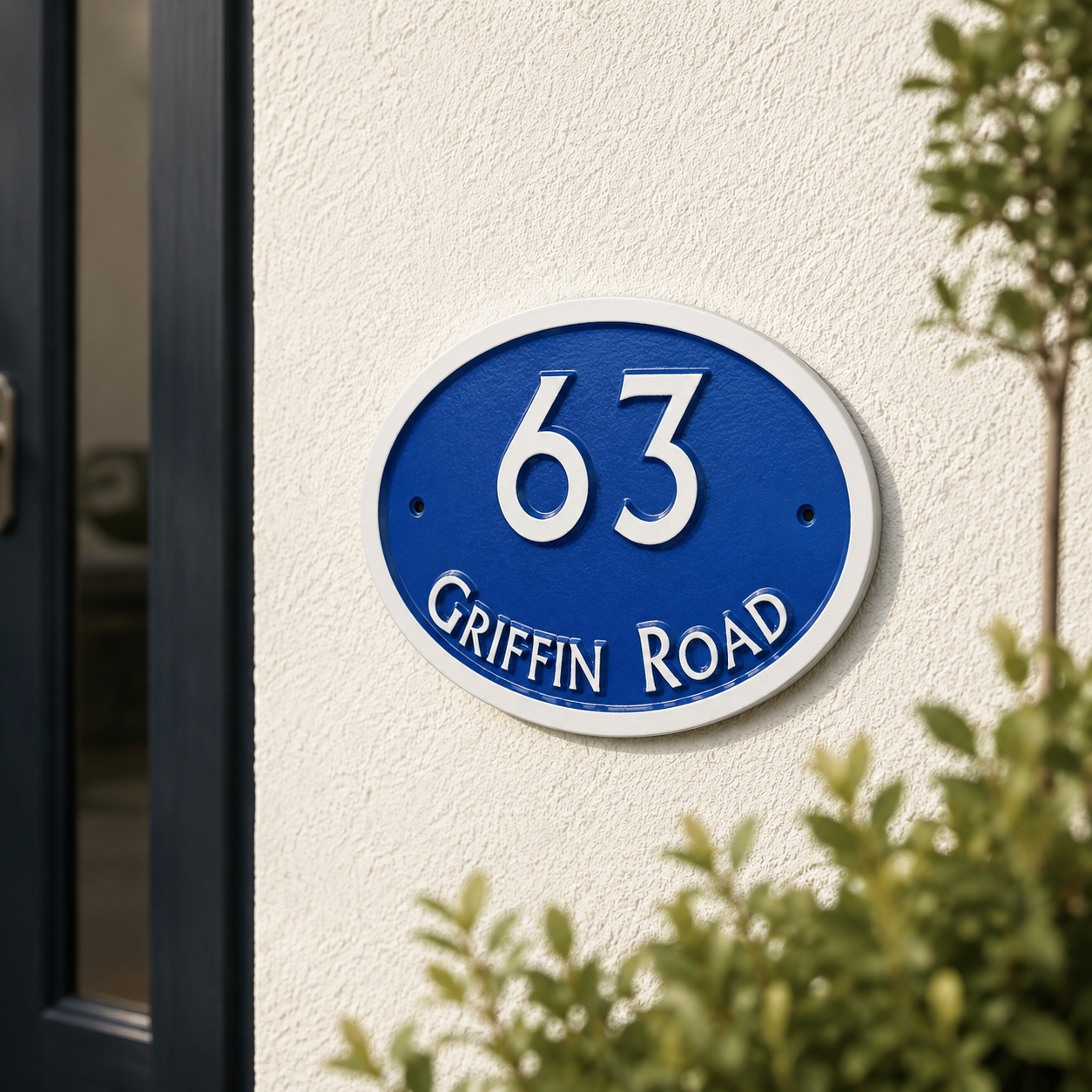 An oval blue and white house number sign reading '63 GRIFFIN ROAD' mounted on a textured white stucco wall, with green foliage partially framing the scene.