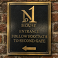 A rectangular black metal house entrance sign with gold lettering and border, mounted on a red-brown brick wall. The sign reads 'N1 HOUSE ENTRANCE FOLLOW FOOTPATH TO SECOND GATE' with a left-pointing arrow.