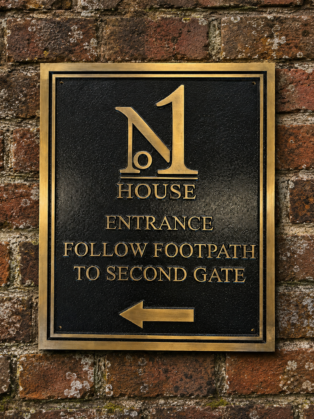 A rectangular black metal house entrance sign with gold lettering and border, mounted on a red-brown brick wall. The sign reads 'N1 HOUSE ENTRANCE FOLLOW FOOTPATH TO SECOND GATE' with a left-pointing arrow.