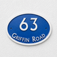 An oval blue house number sign reading '63 GRIFFIN ROAD' in raised white lettering, mounted against a textured white wall.
