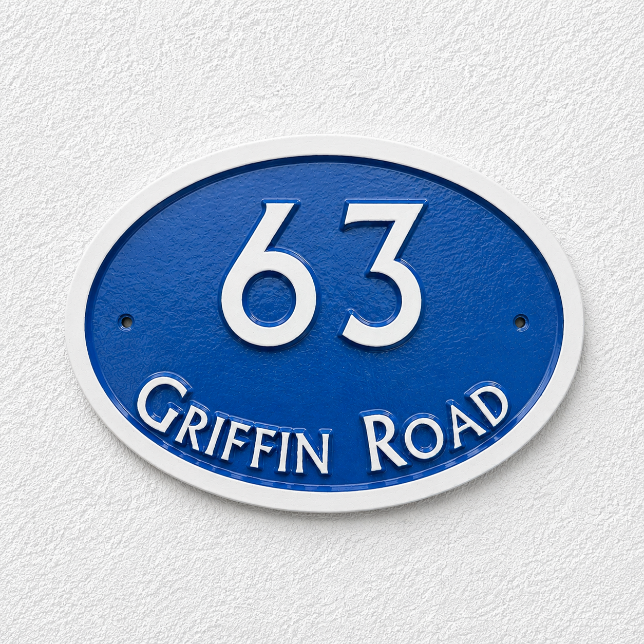 An oval blue house number sign reading '63 GRIFFIN ROAD' in raised white lettering, mounted against a textured white wall.