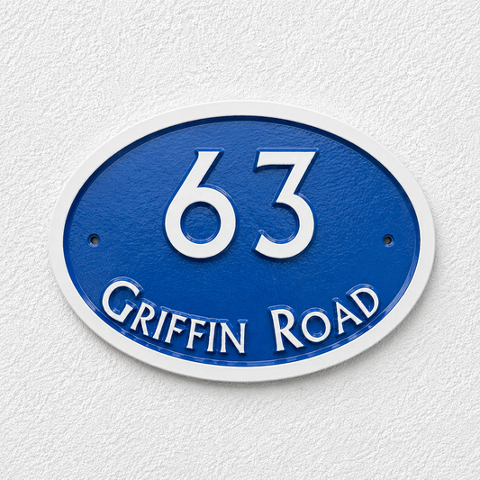 An oval blue house number sign reading '63 GRIFFIN ROAD' in raised white lettering, mounted against a textured white wall.