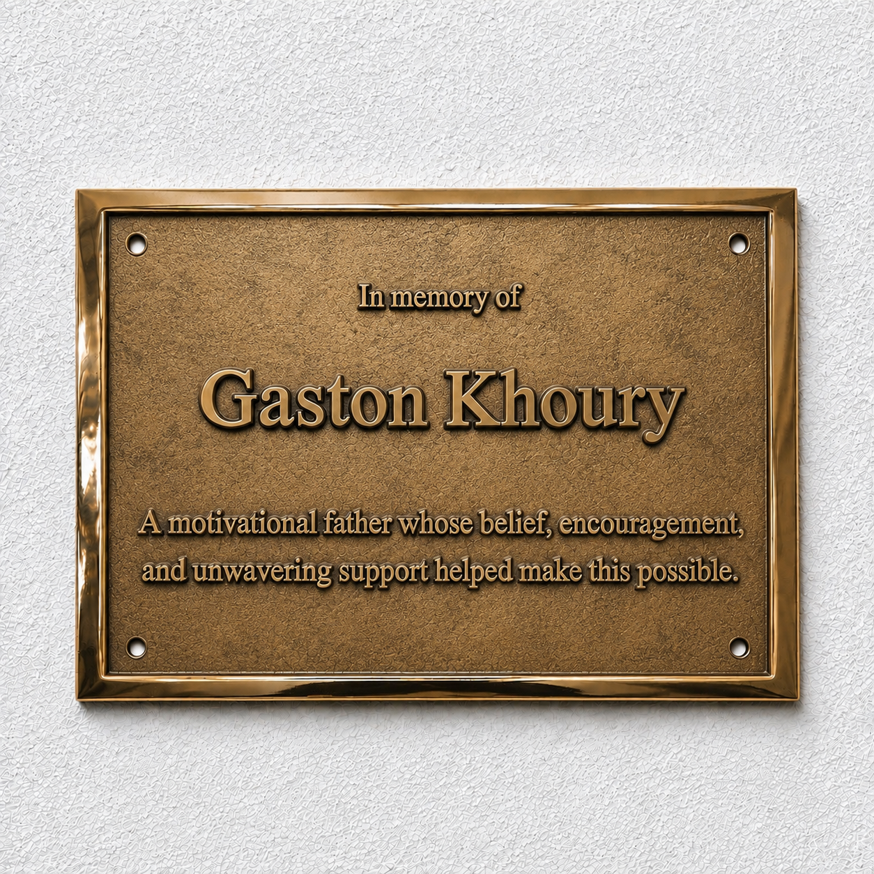 A rectangular bronze memorial plaque mounted on a textured white wall. The plaque features raised text reading 'In memory of Gaston Khoury' and a shorter inscription below, with a polished metallic border and four screw holes.