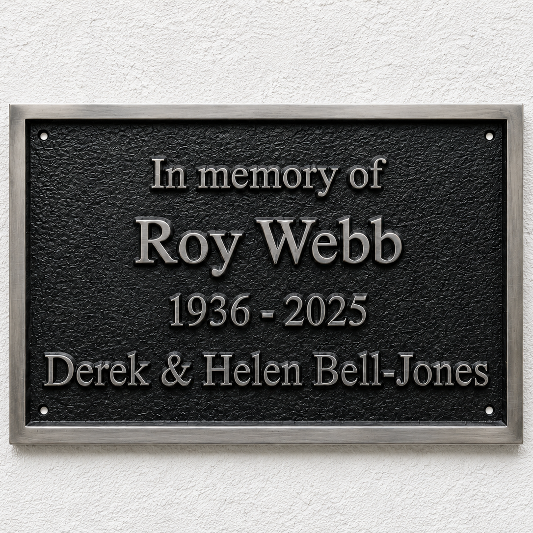 A rectangular black memorial plaque with a brushed silver border, mounted on a white textured wall. The plaque features raised silver lettering that reads 'In memory of Roy Webb 1936 - 2025 Derek & Helen Bell-Jones'.