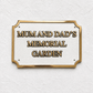 A rectangular memorial garden sign with a gold border and raised gold lettering on a textured white background, mounted on a white wall.