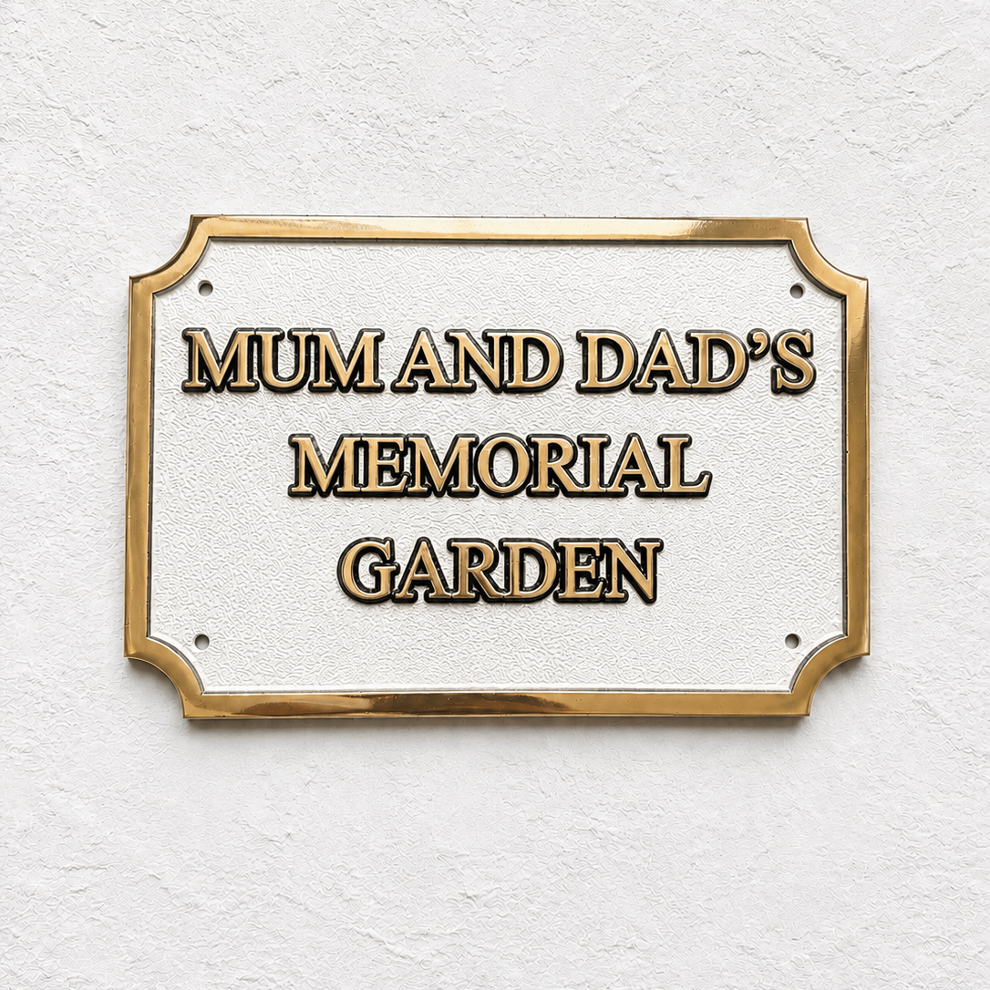 A rectangular memorial garden sign with a gold border and raised gold lettering on a textured white background, mounted on a white wall.