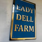 Blue sign with gold text 'LADY DELL FARM' on a white wall