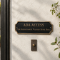 ADA Access doorbell sign mounted on a textured white stucco wall above a dark metal doorbell, with green foliage on the right side.