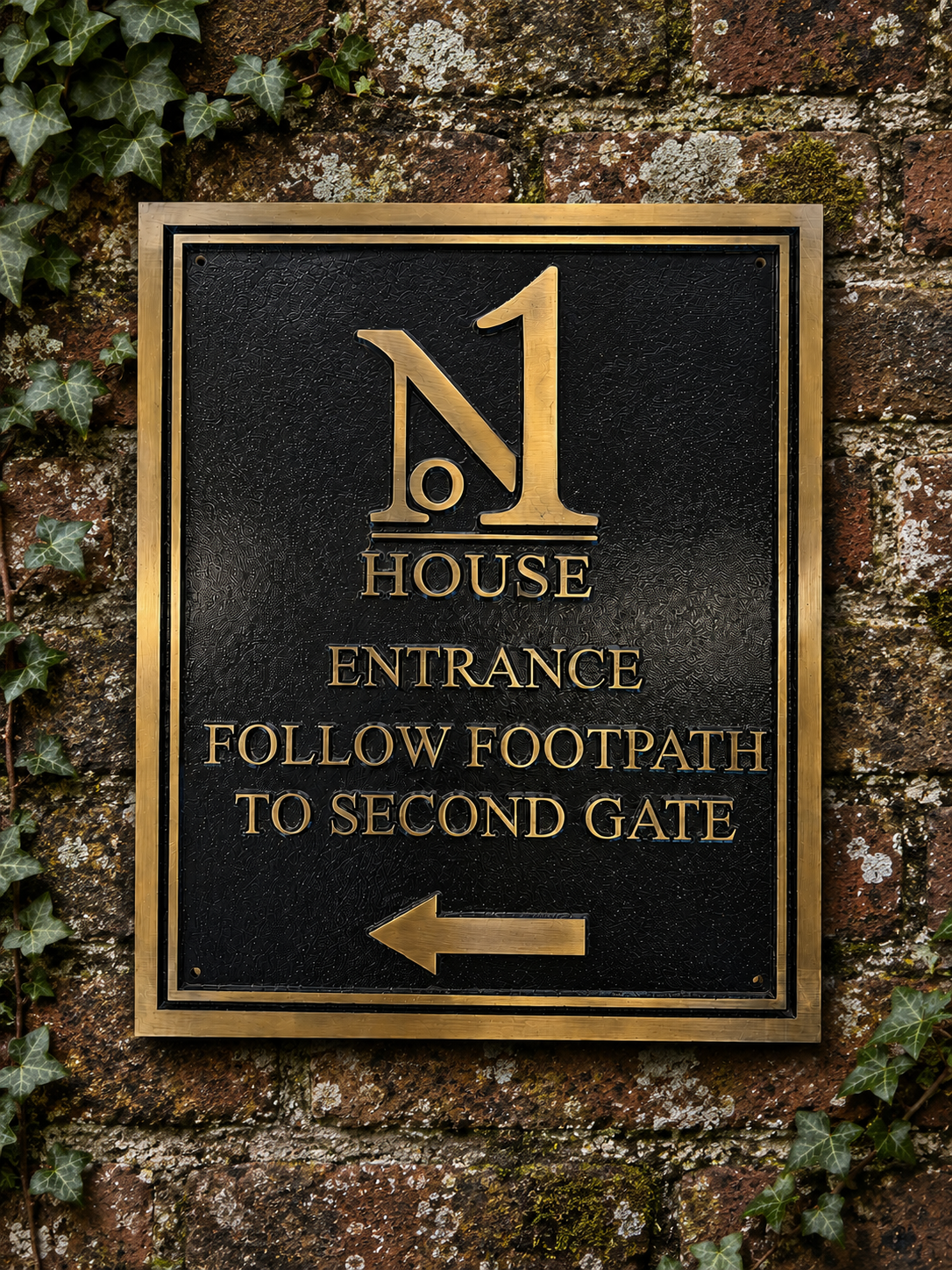A rectangular black metal sign with a gold border and gold lettering mounted on a weathered brick wall with ivy. The sign reads 'No 1 House Entrance Follow Footpath to Second Gate' with a left-pointing arrow at the bottom.