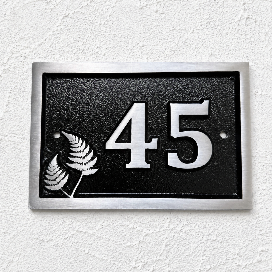 A rectangular black house number plaque with a silver border, featuring the number 45 in raised silver lettering and two silver fern leaves on the left, mounted on a textured white wall.