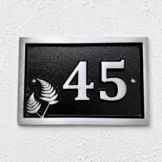 A rectangular black house number plaque with a silver border, featuring the number 45 in raised silver lettering and two silver fern leaves on the left, mounted on a textured white wall.