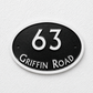 An oval black house number plaque with a white border, featuring the number 63 in white and the text 'GRIFFIN ROAD' in white along the bottom, mounted on a white textured wall.