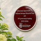 A round dark red commemorative plaque mounted on a textured white wall, with green leaves and pale flowers blurred in the foreground. The plaque reads: 'Generously supported by the National Heritage Memorial Fund and UK government funding 2023-2026' and includes the National Heritage Memorial Fund logo.
