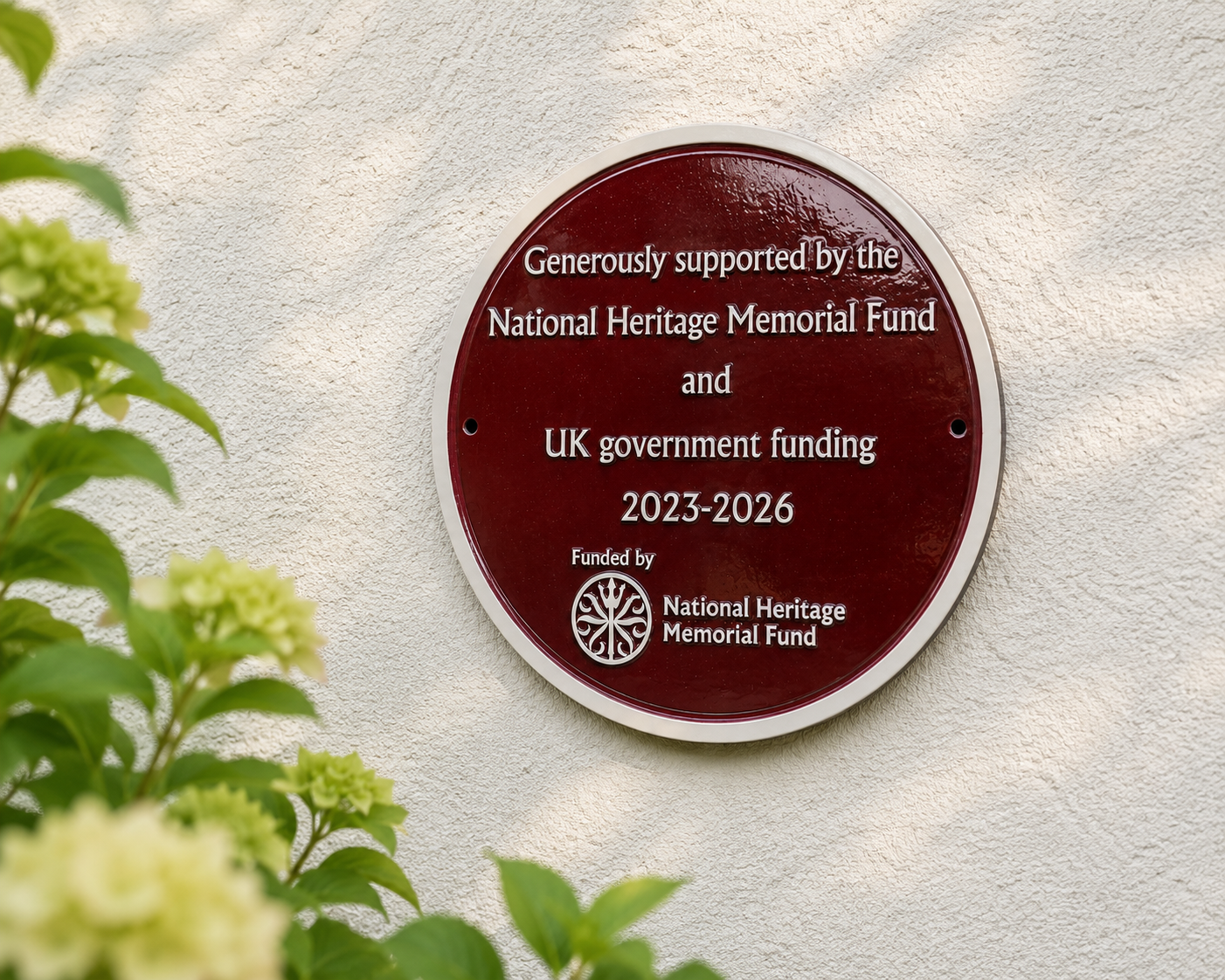 A round dark red commemorative plaque mounted on a textured white wall, with green leaves and pale flowers blurred in the foreground. The plaque reads: 'Generously supported by the National Heritage Memorial Fund and UK government funding 2023-2026' and includes the National Heritage Memorial Fund logo.