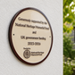 An oval commemorative plaque mounted on a textured white wall, with a blurred outdoor garden and stone building in the background. The plaque is cream-colored with a dark brown border and engraved text reading 'Generously supported by the National Heritage Memorial Fund and UK government funding 2023-2026' and 'Funded by National Heritage Memorial Fund' with a circular emblem.