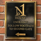 A rectangular black metal house entrance sign with a gold border, featuring a large gold 'N1' logo, the words 'HOUSE ENTRANCE', 'FOLLOW FOOTPATH TO SECOND GATE', and a left-pointing arrow, mounted on a red brick wall.