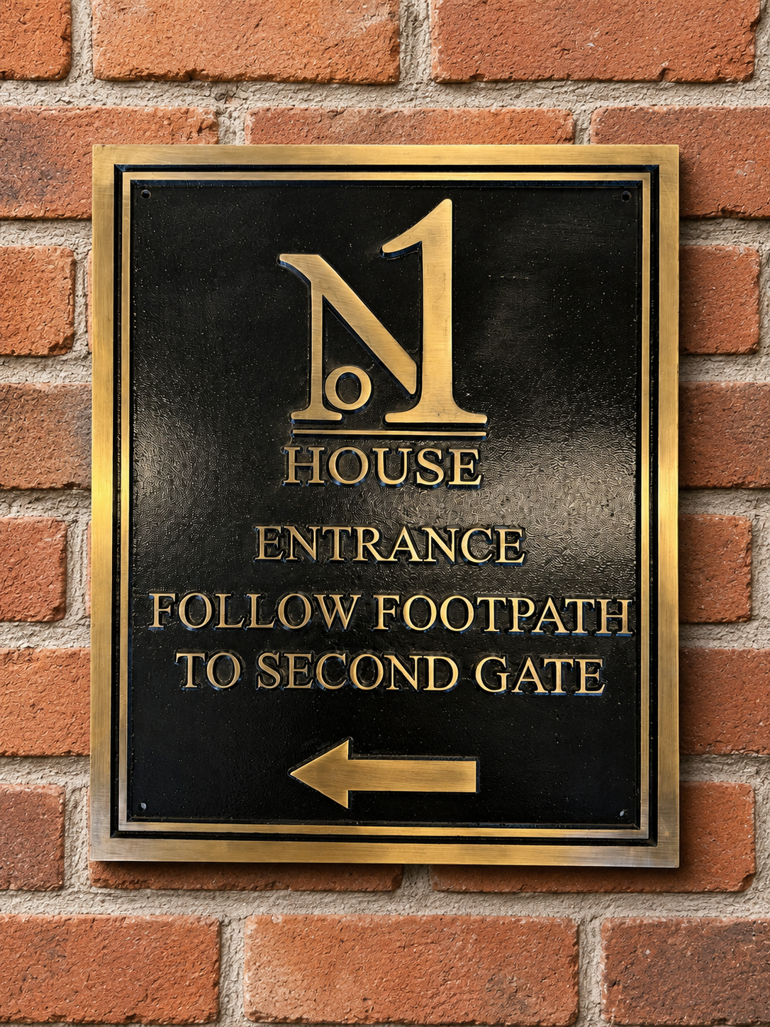 A rectangular black metal house entrance sign with a gold border, featuring a large gold 'N1' logo, the words 'HOUSE ENTRANCE', 'FOLLOW FOOTPATH TO SECOND GATE', and a left-pointing arrow, mounted on a red brick wall.
