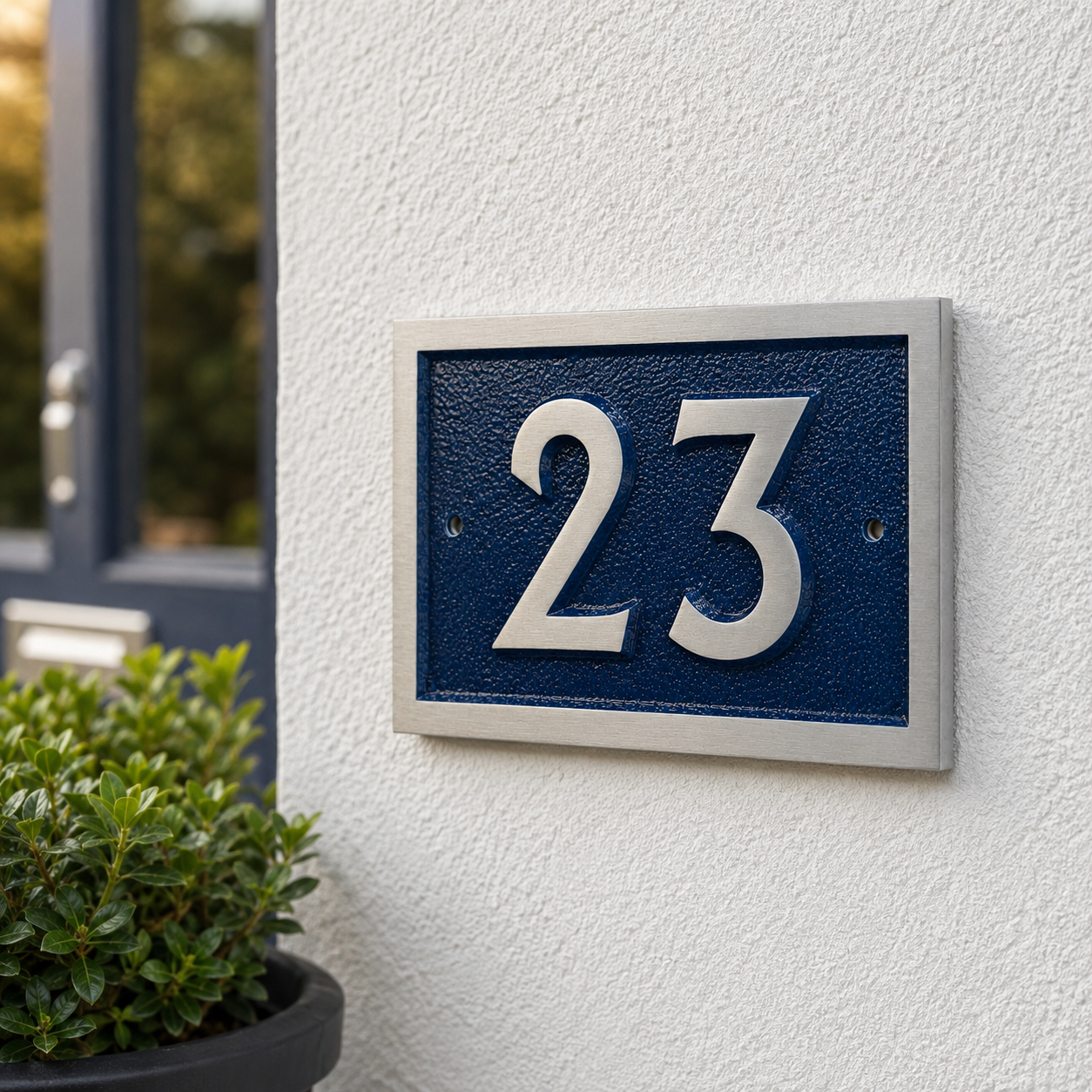 A modern house number plaque featuring the number '23' in raised silver lettering on a textured dark blue background, mounted on a white stucco wall next to a potted green plant.