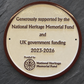 A round bronze and cream metal commemorative plaque mounted on a dark slate background, featuring engraved text for the National Heritage Memorial Fund and UK government funding, 2023-2026.