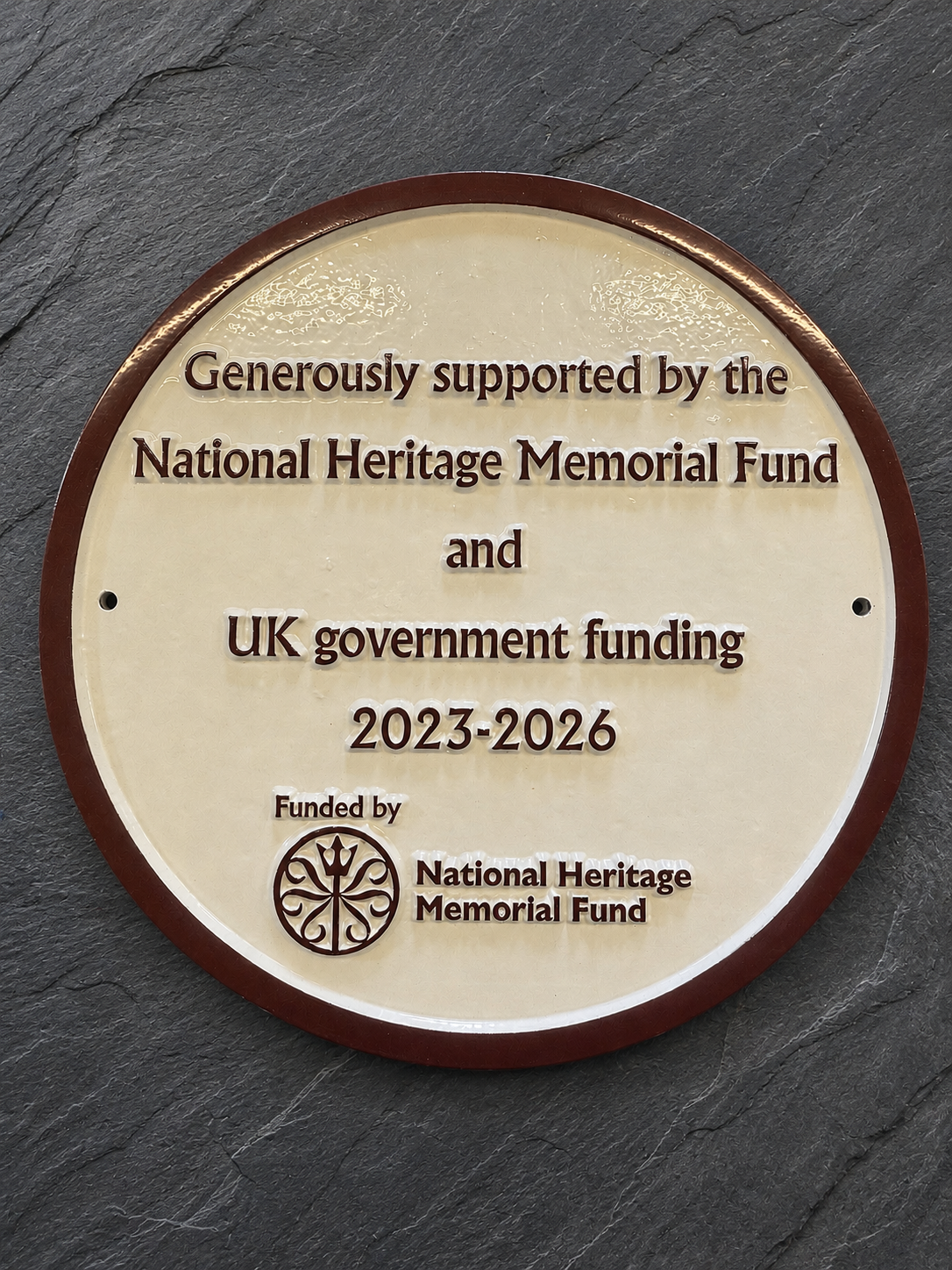 A round bronze and cream metal commemorative plaque mounted on a dark slate background, featuring engraved text for the National Heritage Memorial Fund and UK government funding, 2023-2026.