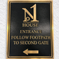 A rectangular black metal entrance sign with a gold border, featuring the text 'No 1 House Entrance Follow Footpath to Second Gate' and a left-pointing arrow, mounted on a white textured wall.