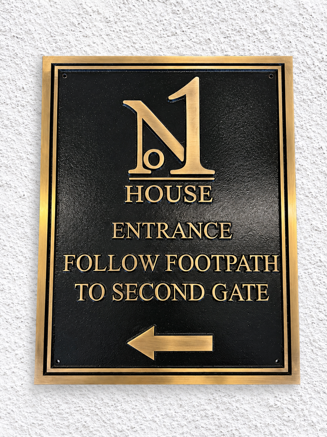 A rectangular black metal entrance sign with a gold border, featuring the text 'No 1 House Entrance Follow Footpath to Second Gate' and a left-pointing arrow, mounted on a white textured wall.