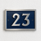 A rectangular house number plaque with raised silver numerals '23' on a textured dark blue background, mounted on a white textured wall.