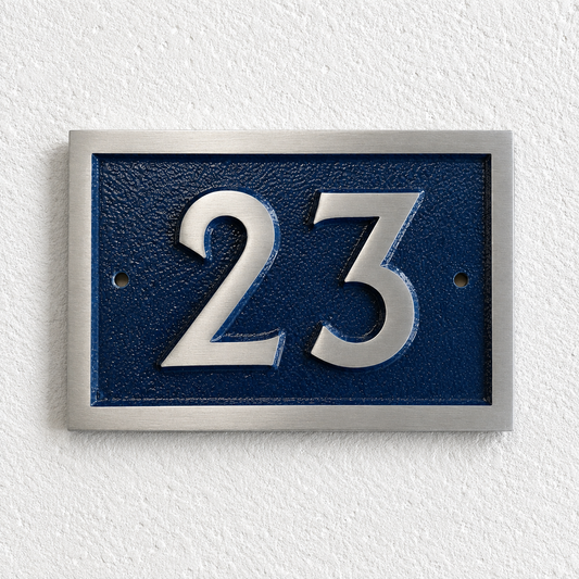 A rectangular house number plaque with raised silver numerals '23' on a textured dark blue background, mounted on a white textured wall.