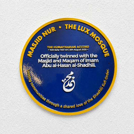 A round blue and yellow plaque mounted on a white textured wall, featuring text for Masjid Nur and The Lux Mosque with a white Arabic calligraphy symbol in the center.