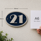An oval house number plaque shown mounted on a textured wall, with dimension lines indicating 160mm width and 110mm height. A hand holds a white card labeled A6 next to it for scale.