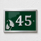 A rectangular green house number plaque with silver numbers '45' and silver fern motifs, mounted on a white textured wall.