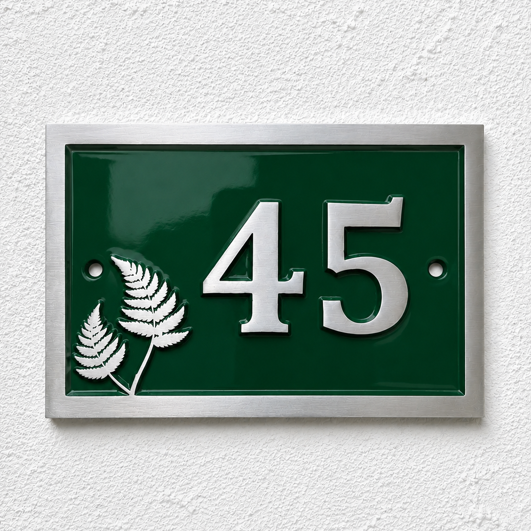 A rectangular green house number plaque with silver numbers '45' and silver fern motifs, mounted on a white textured wall.
