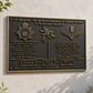 A bronze military commemorative plaque mounted on a white stucco wall, with green foliage visible at the left and right edges. The plaque features raised gold-toned insignia and text for the 12th Battalion - The Devonshire Regiment and 6th Airborne Division in Normandy.