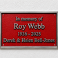 A rectangular red memorial plaque with a silver metal frame, mounted on a white textured wall. The plaque features raised silver text reading 'In memory of Roy Webb 1936 - 2025 Derek & Helen Bell-Jones'.