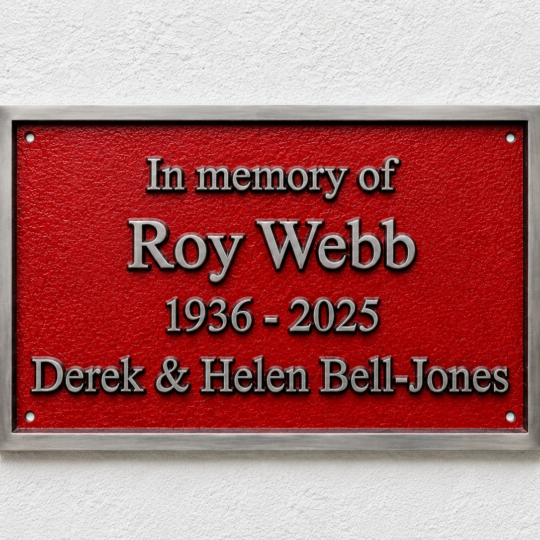 A rectangular red memorial plaque with a silver metal frame, mounted on a white textured wall. The plaque features raised silver text reading 'In memory of Roy Webb 1936 - 2025 Derek & Helen Bell-Jones'.