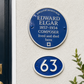 A blue circular English Heritage plaque for composer Edward Elgar mounted on a textured white wall, with a smaller blue oval house number 63 plaque below and part of a dark door and green foliage visible at the edges.