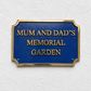 A rectangular blue memorial garden sign with gold lettering reading 'MUM AND DAD'S MEMORIAL GARDEN' and a gold border, mounted on a textured white wall.
